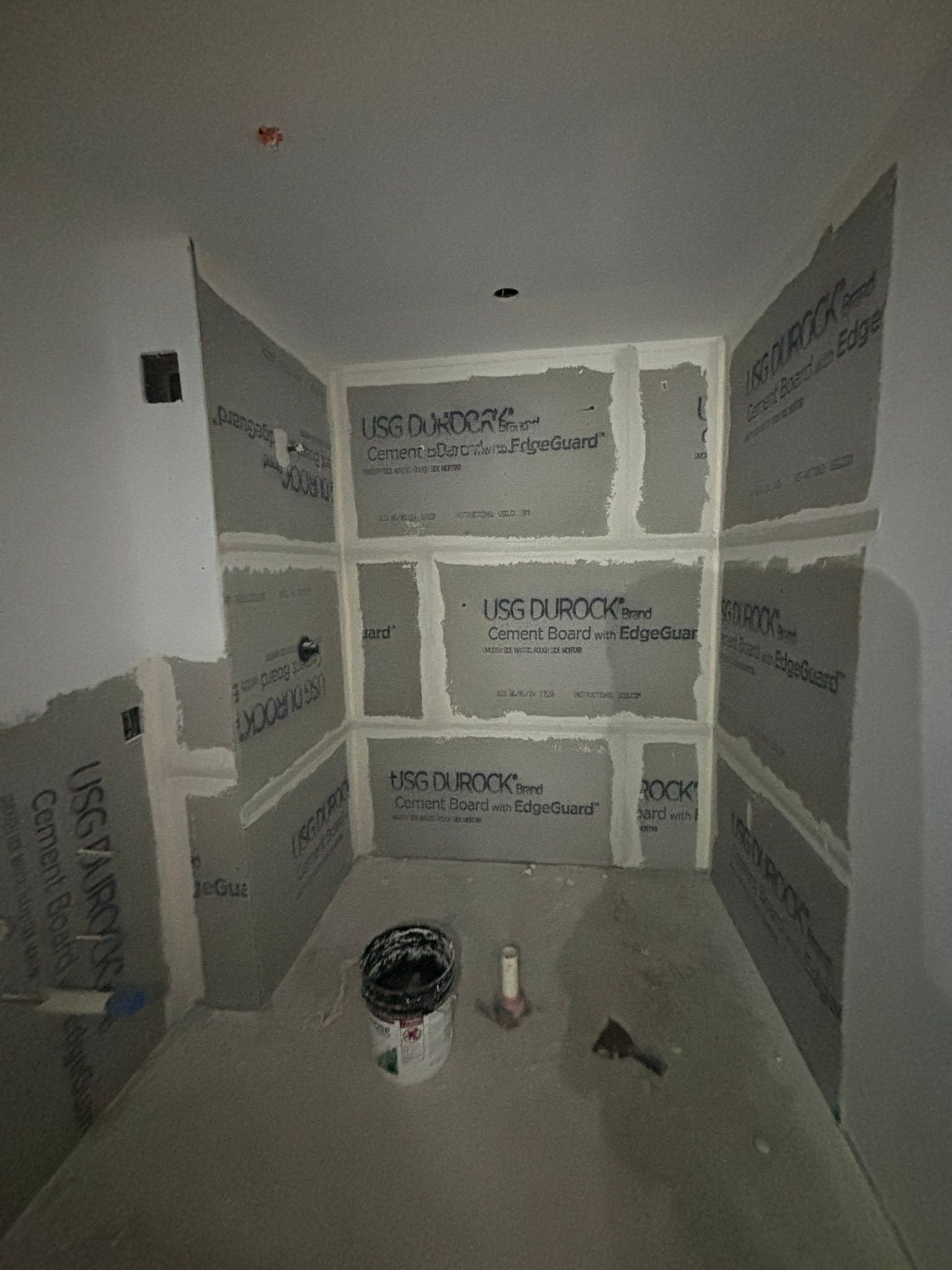 Bathroom under construction, gray cement board walls with white joint compound, bucket and tools on floor.