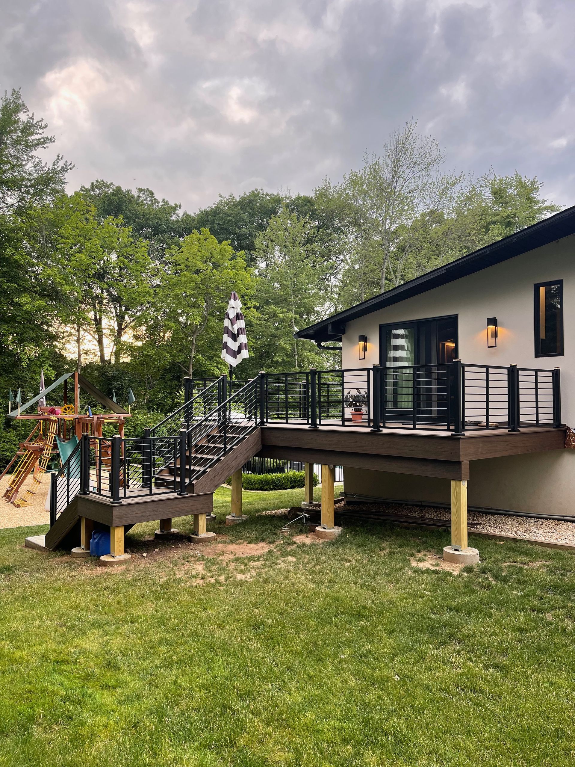A home with a wooden deck, black railings, and an umbrella, surrounded by green grass and trees.
