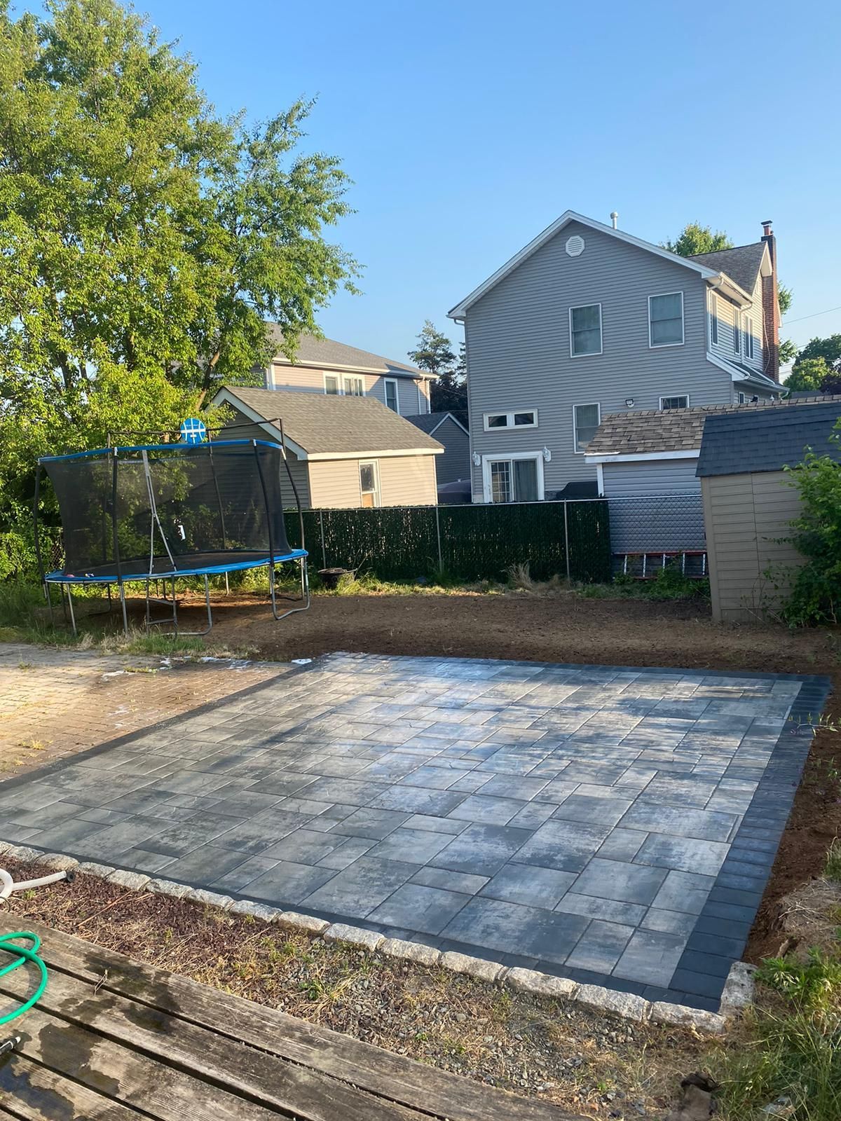 Backyard with a gray paver patio bordered by black pavers, trampoline, and houses.