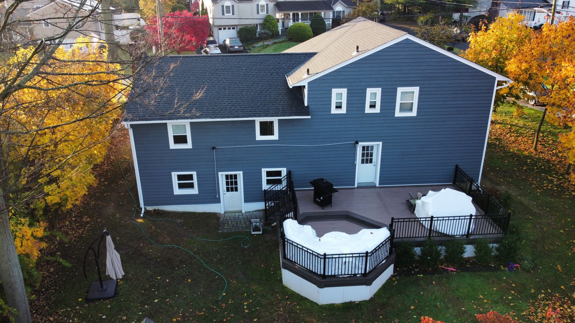 Back of a two-story blue house with a deck and hot tub, surrounded by autumn trees with colorful foliage.
