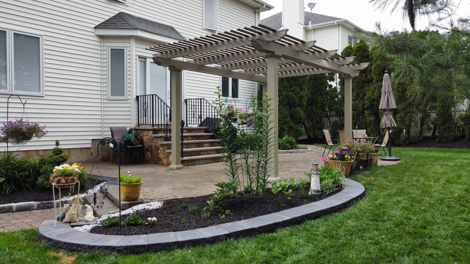 Backyard patio with pergola, steps, and flower beds, next to a white house with lawn.