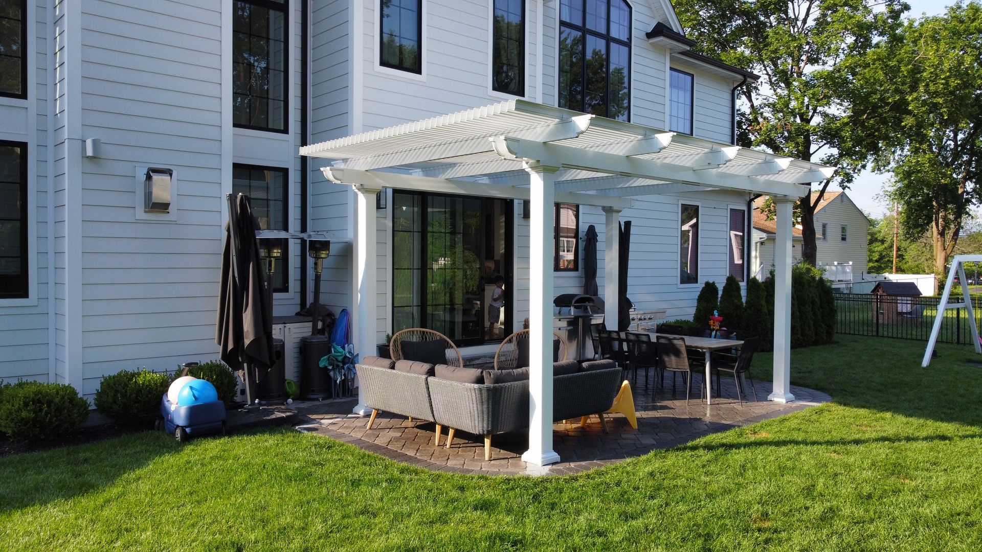 White pergola over a patio with outdoor furniture, adjacent to a white house with black window frames.