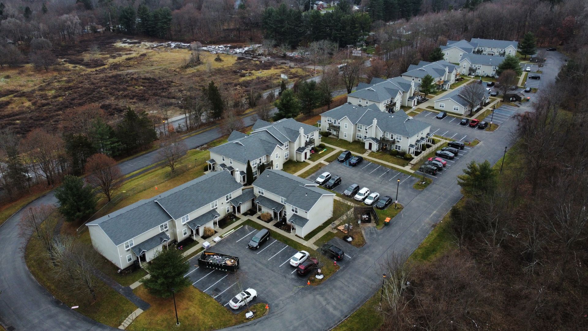Aerial view of apartment complex with parking, surrounded by trees and a field.