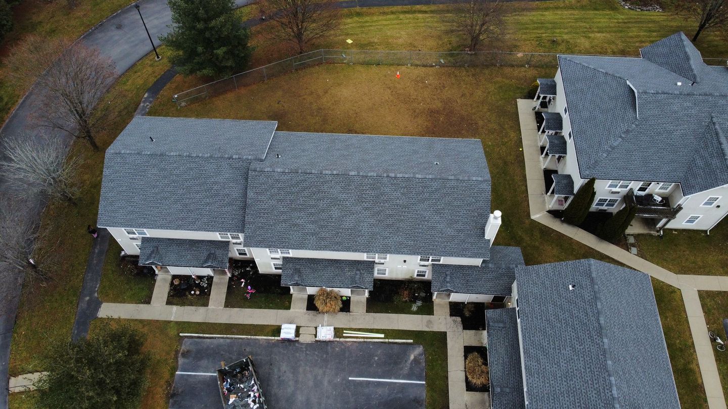 Aerial view of gray-roofed townhouses with small yards, concrete walkways, and a curved road in a grassy area.
