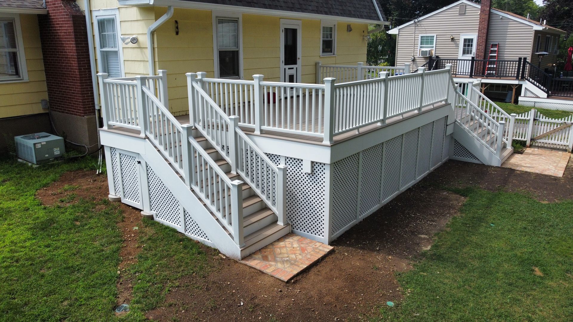 White wooden deck with stairs attached to a yellow house and a gravel path.