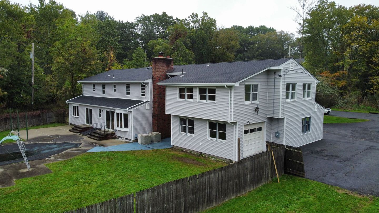 Gray two-story house with black roof, chimney, garage, and a fenced yard with green grass and trees.