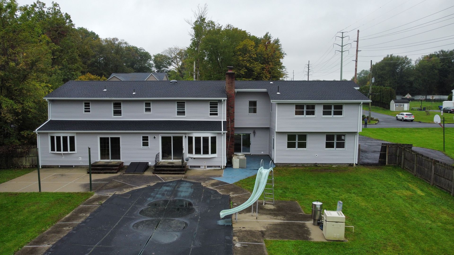 Rear exterior view of a two-story gray house with a covered pool and lawn. Trees in background.