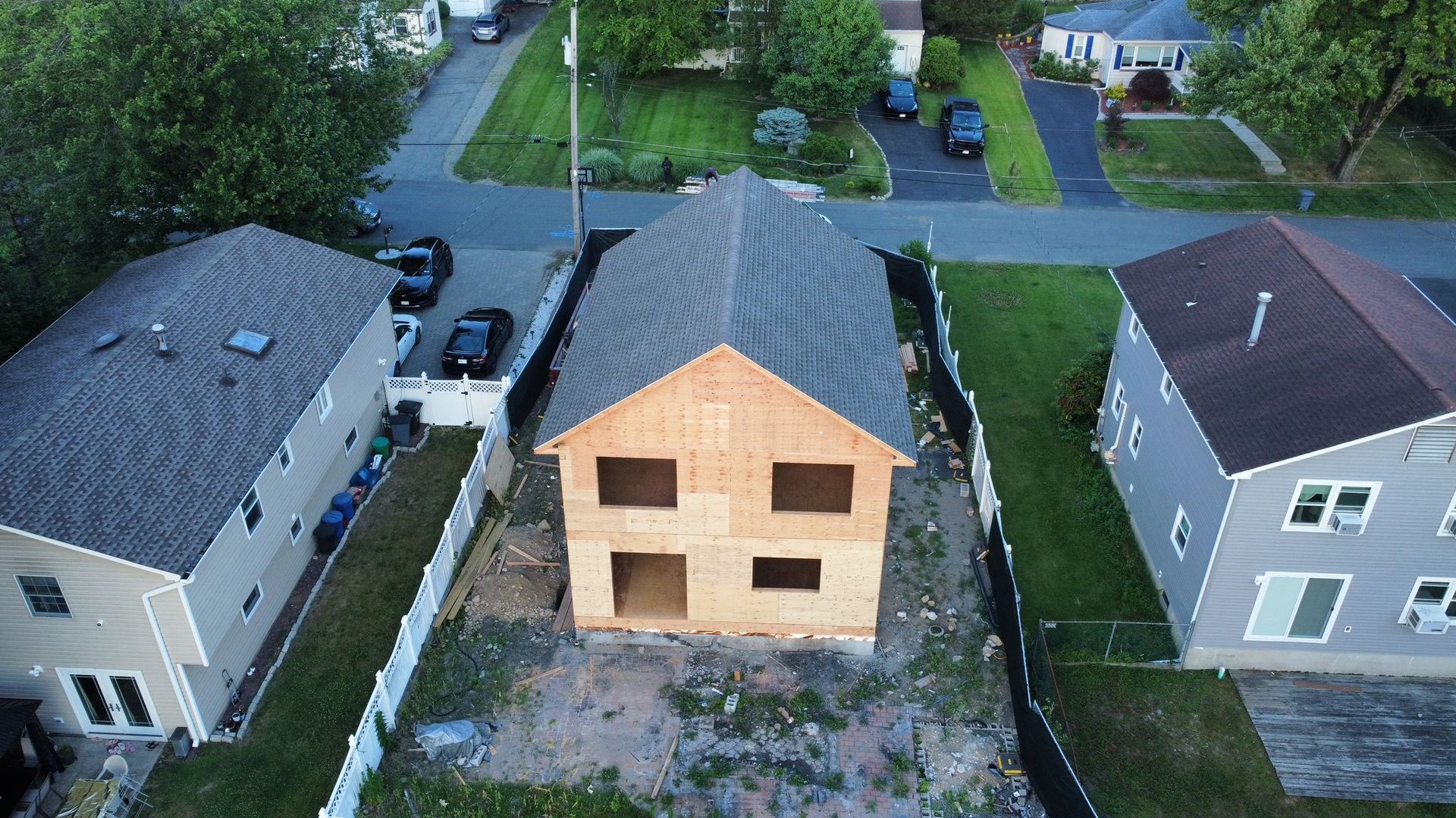 Aerial view of a two-story house under construction, framed between finished houses with parked cars.