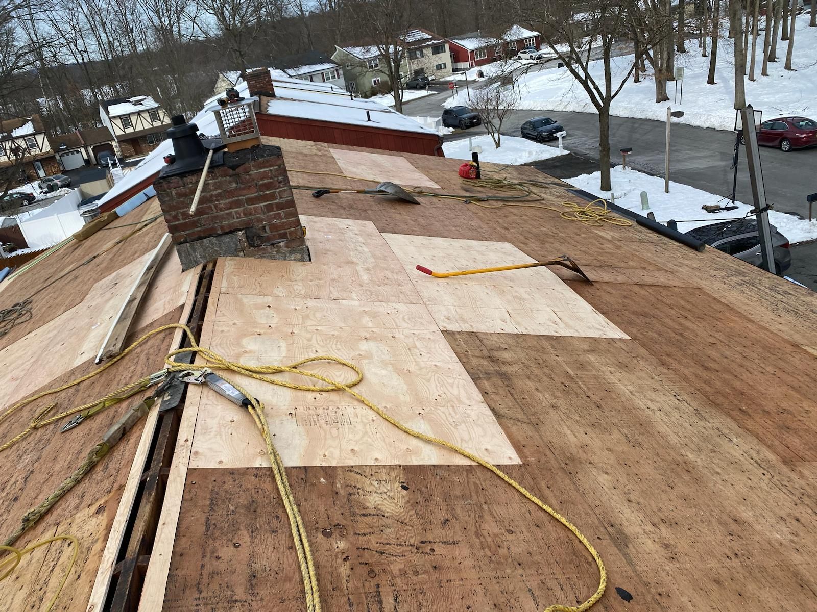 Rooftop with exposed plywood, safety harness, chimney, and winter surroundings.