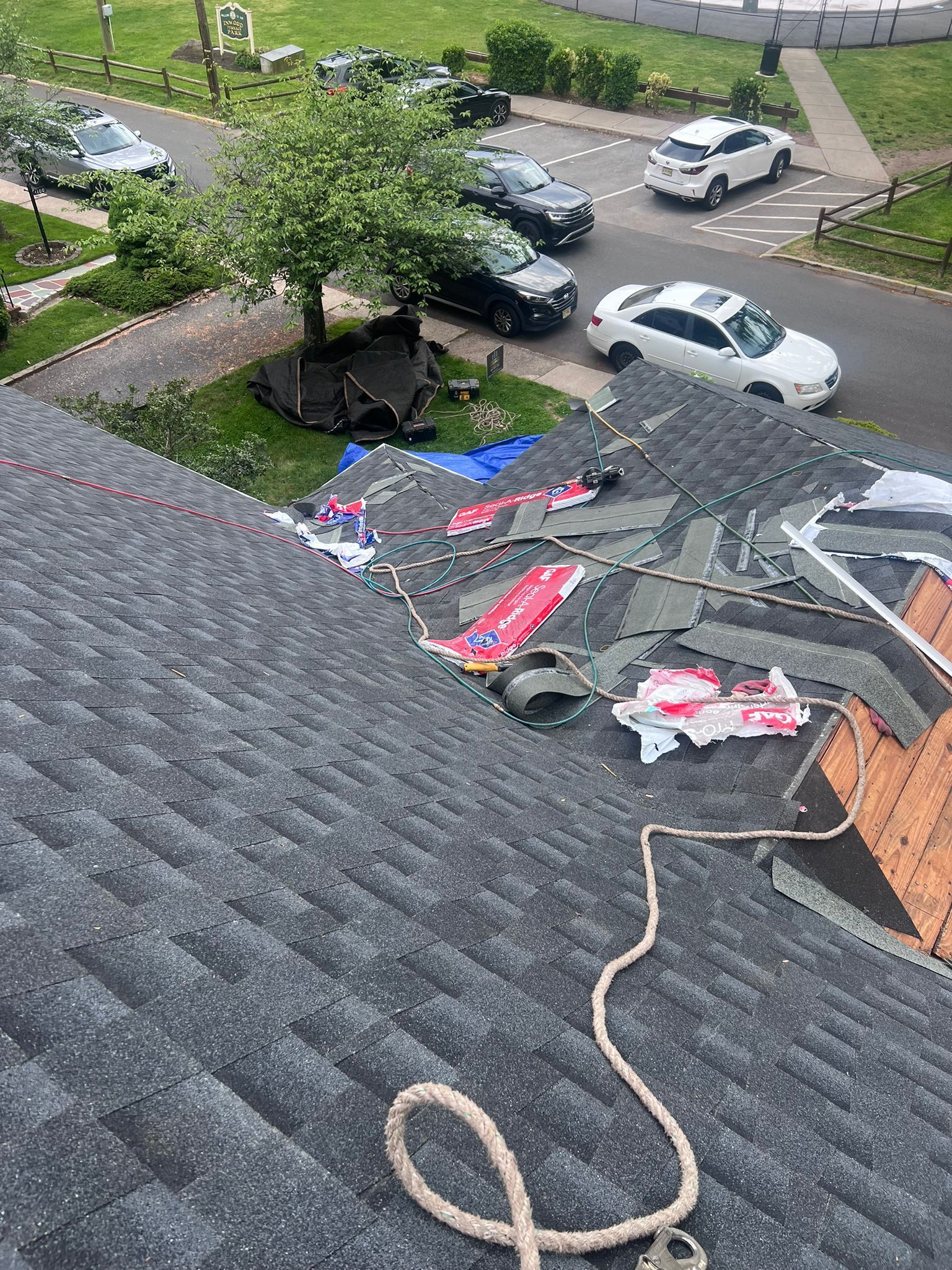 Rooftop with damaged shingles. Debris scattered, rope hangs down. Cars and trees in background.