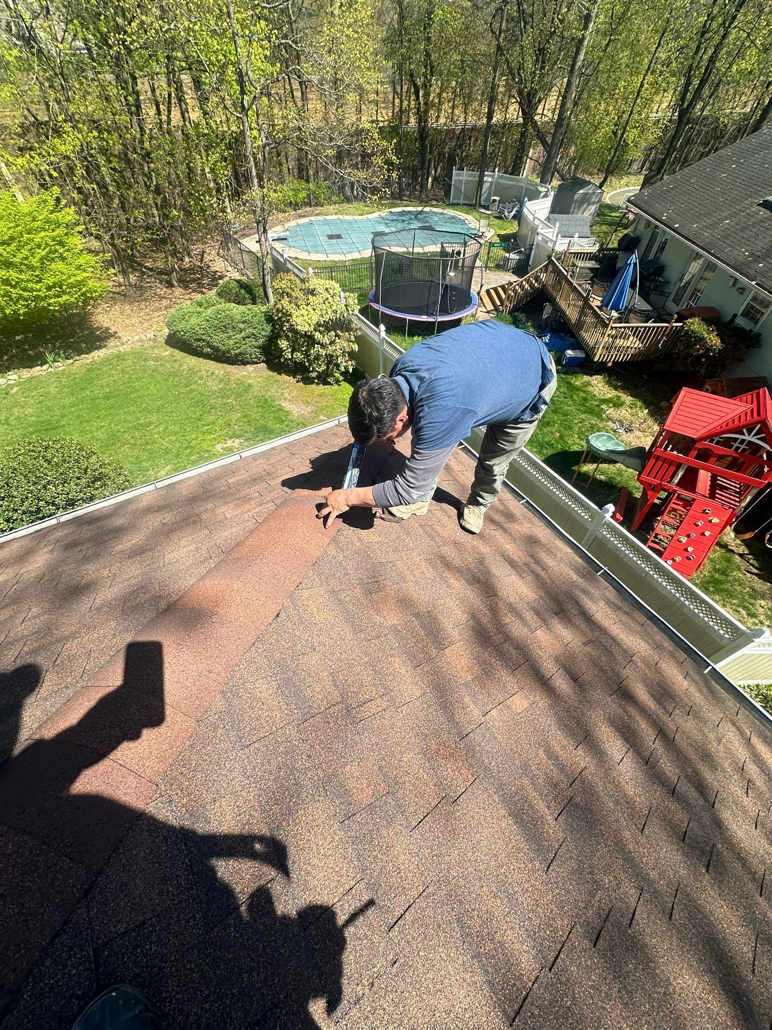 Person on a roof repairs a dark, weathered shingle roof. Green yard, pool, and trees in background.