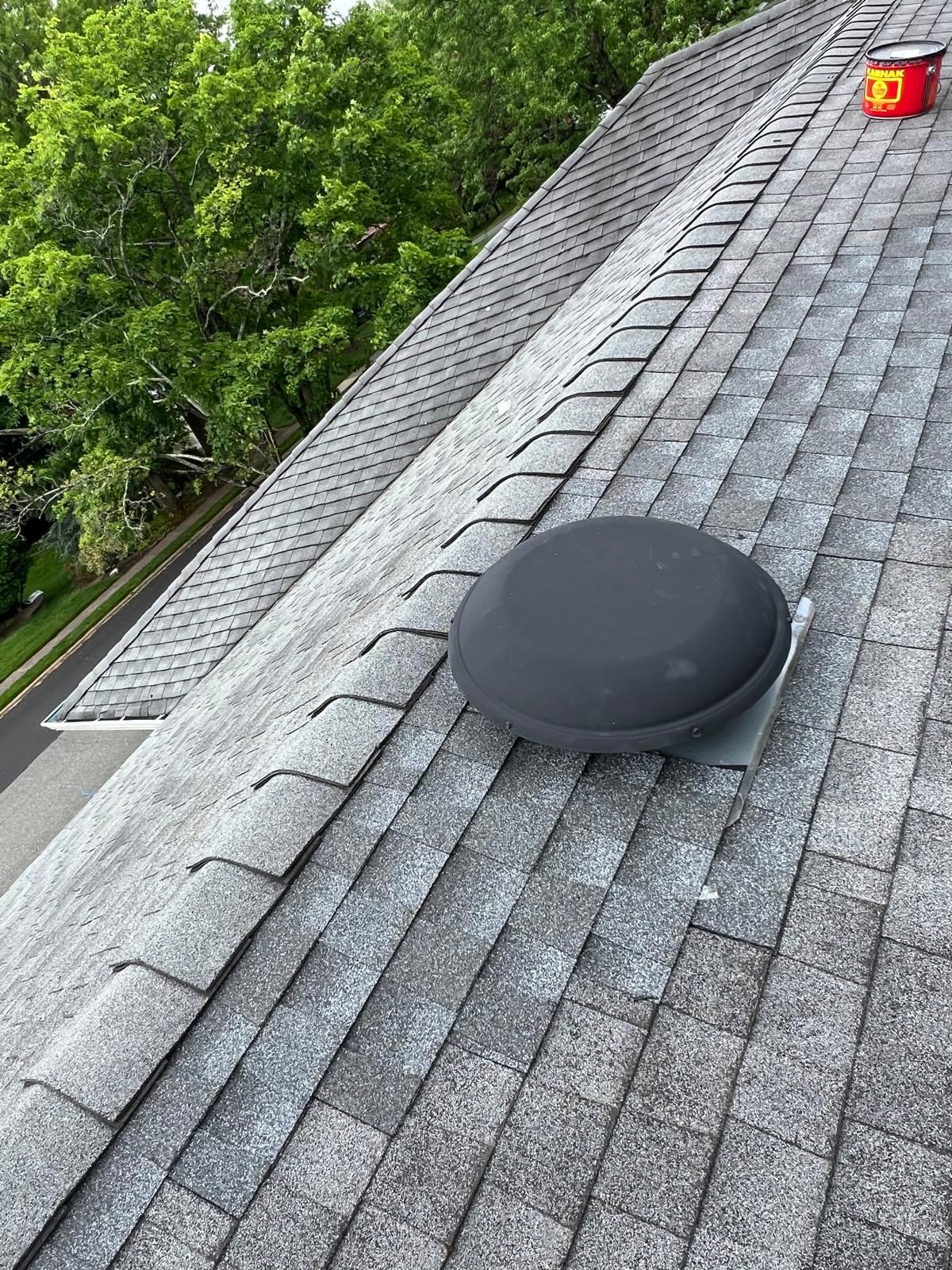 A gray shingle roof with a round, black vent in the center, a red object on the right side.