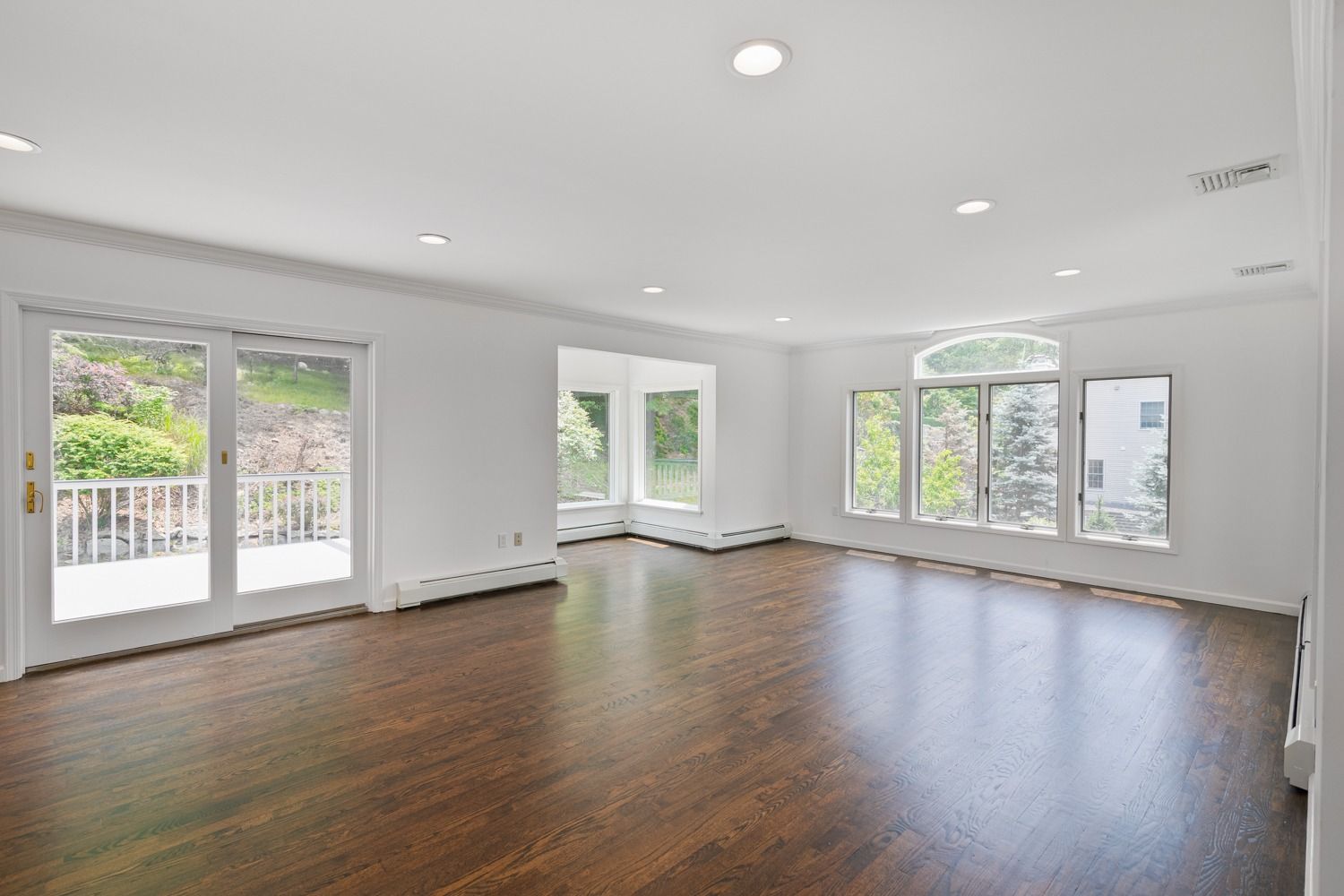 Empty, bright living room with dark hardwood floors, windows, and door to a balcony.