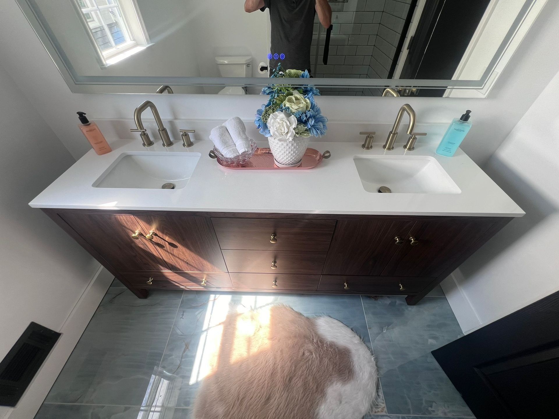 Bathroom with double vanity, white countertop, dark wood cabinets, gold faucets, and a light blue rug.