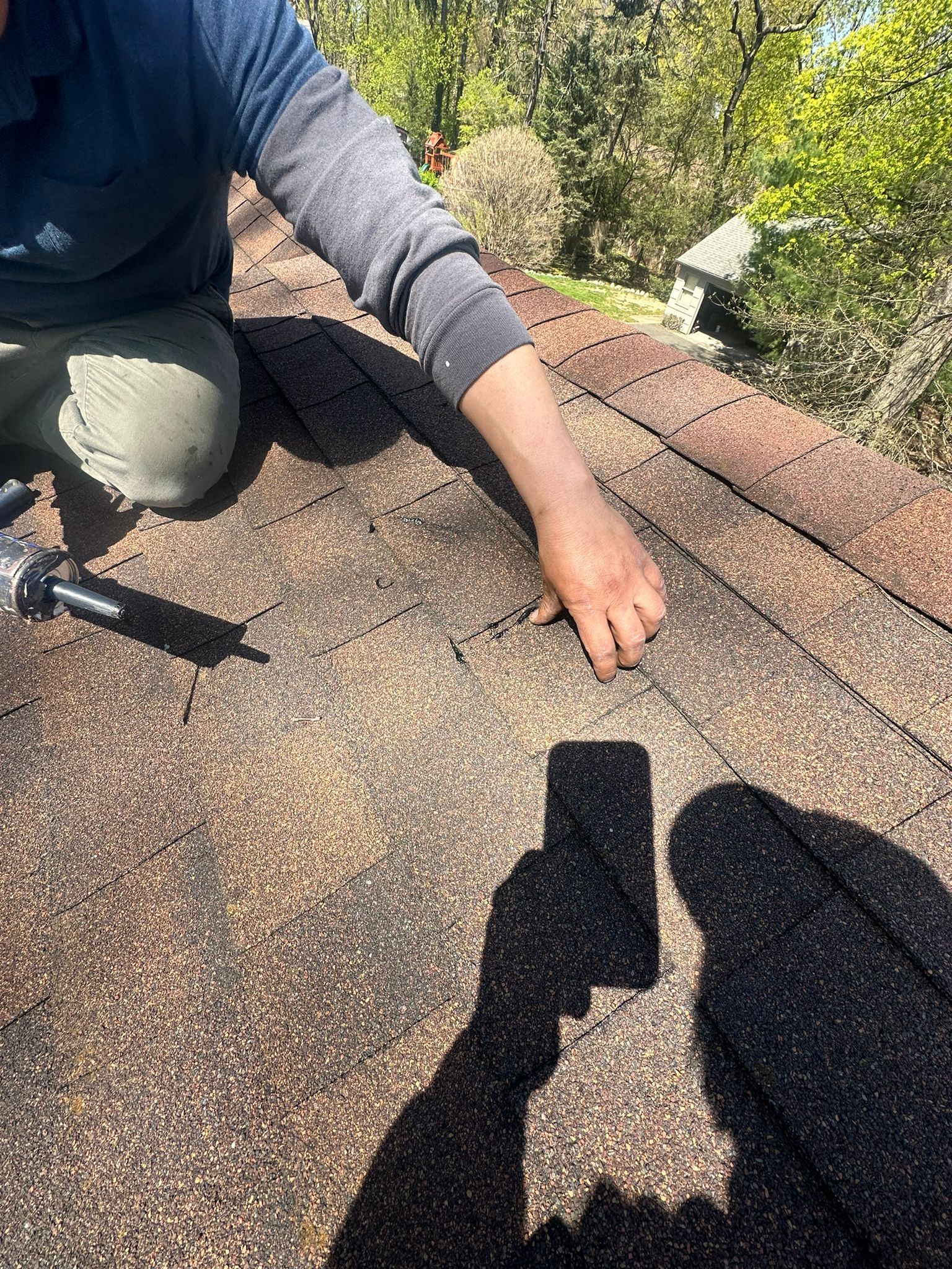 Person on a roof inspecting shingles, hand touching a crack. A cell phone casts a shadow.