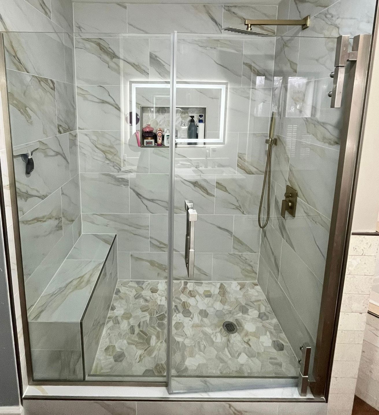 Shower with marble-look tile, glass doors, built-in bench, and pebble-stone floor. Gold fixtures.