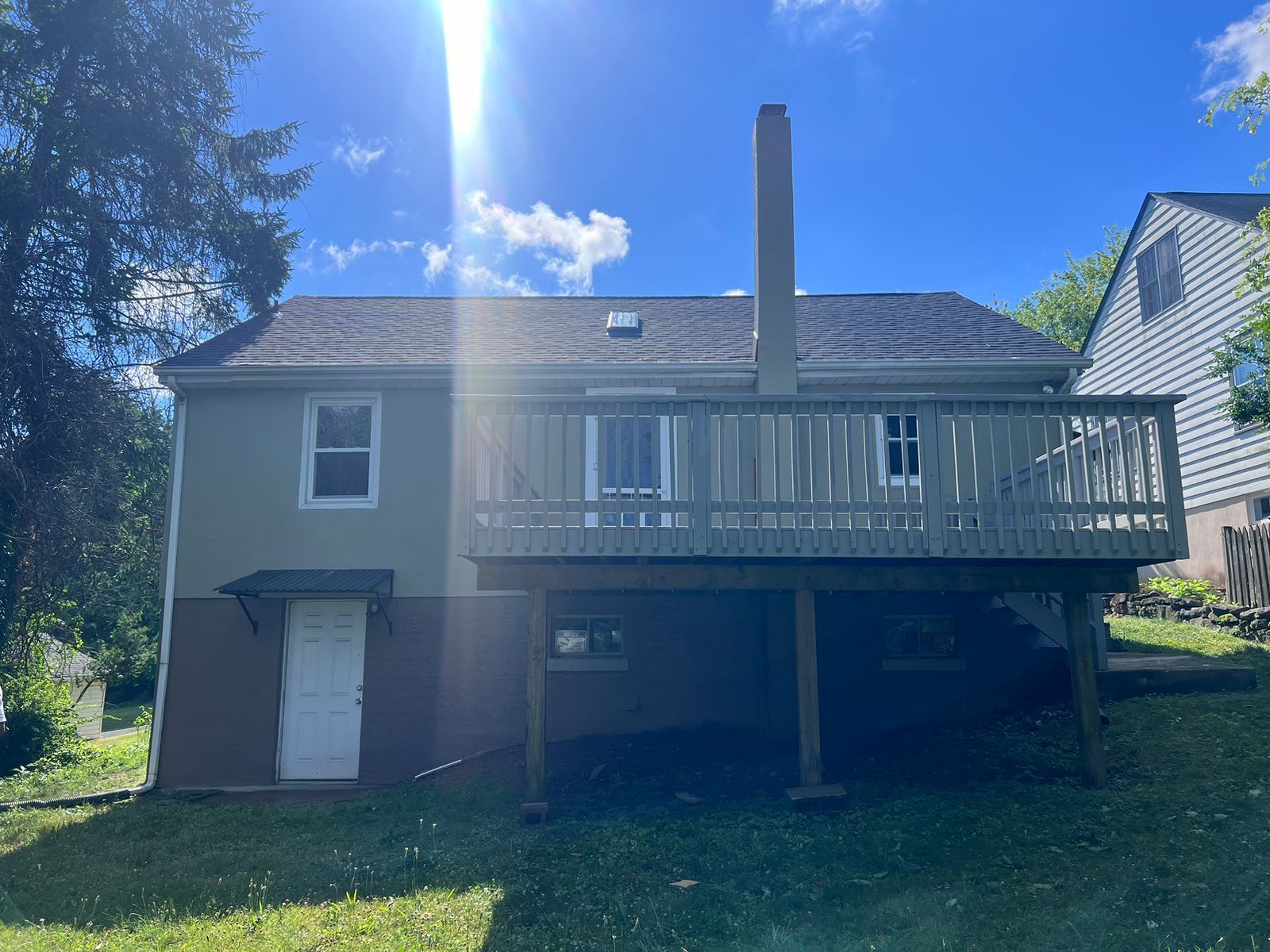 Rear view of a two-story house with a wooden deck and a chimney. Green grass and blue sky are visible.