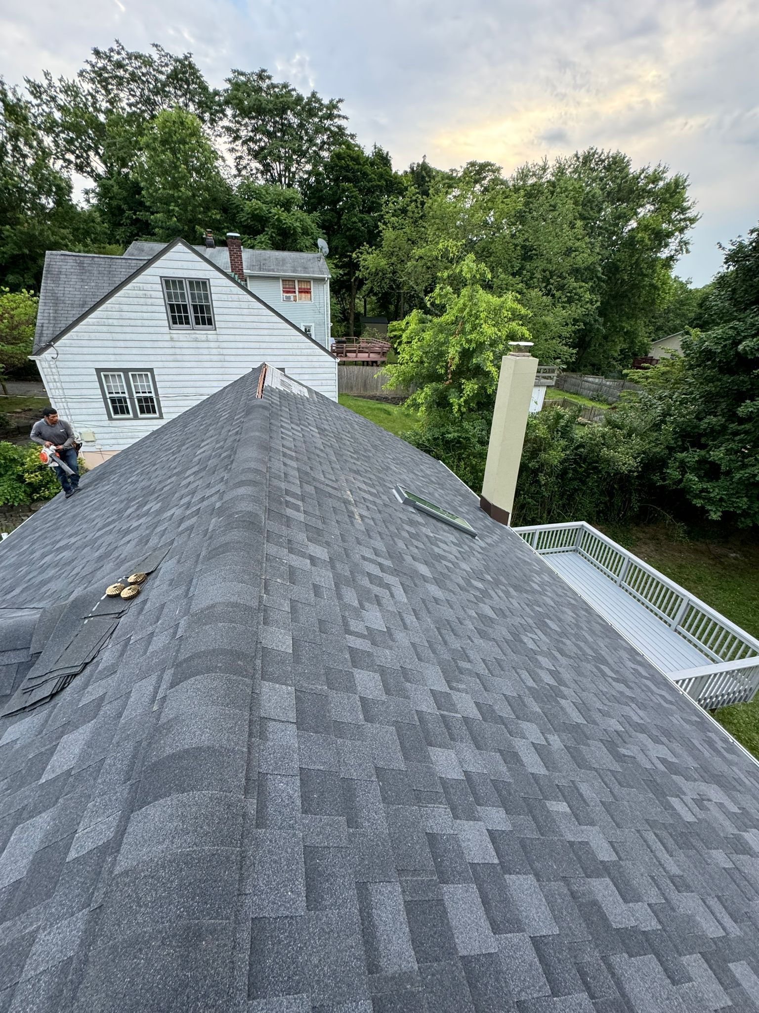 View of a gray shingle roof, chimney, and a white house with trees in the background.