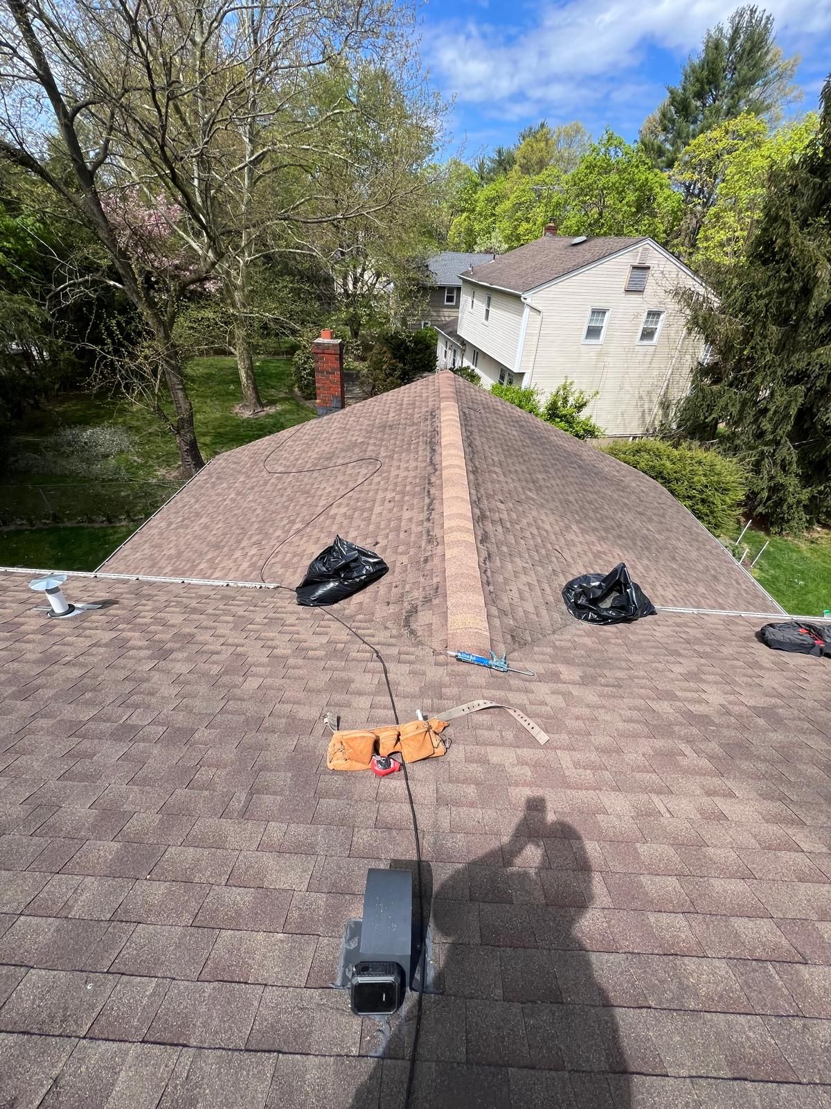 View of a brown shingled roof with a brick chimney. Two black bags and tools on the roof. Bright sunny day.