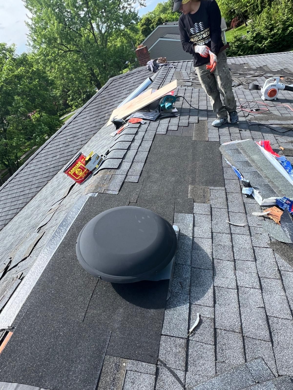 Person on a roof repairing shingles around a large black vent. Sunny outdoor setting.