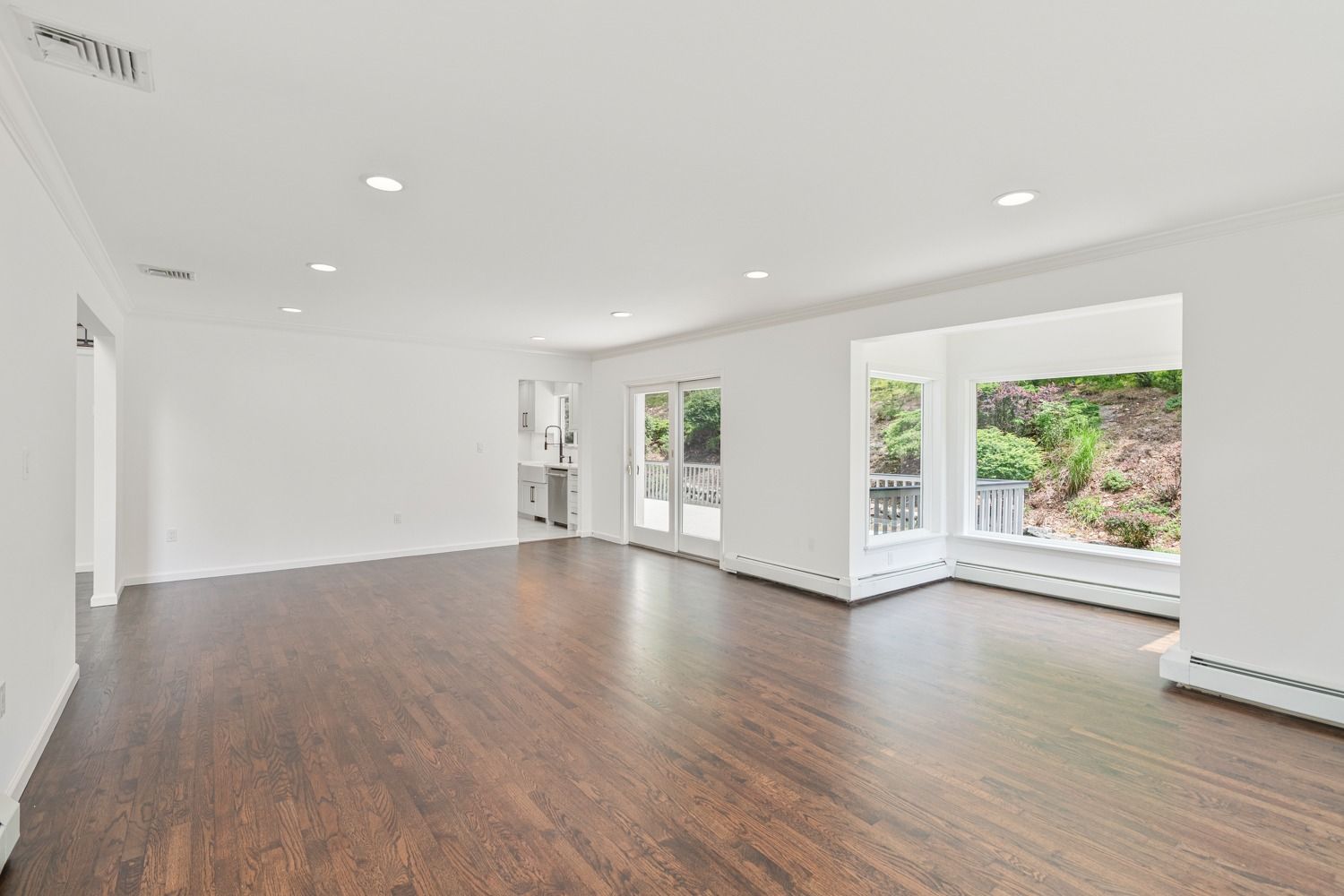 Empty, bright living room with dark hardwood floors, white walls, and large windows with a view.