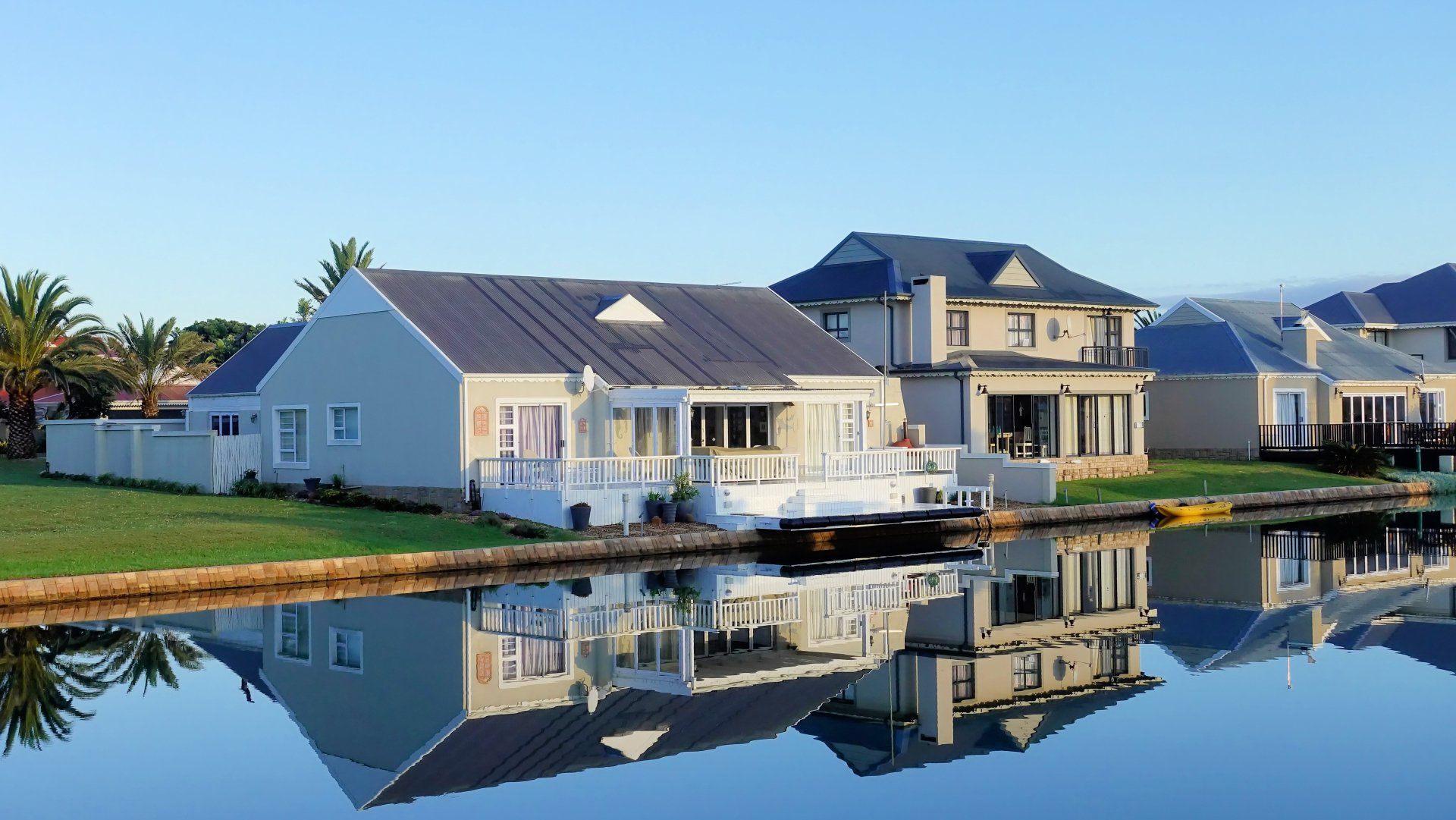 Waterfront houses reflect in calm water under a clear blue sky.