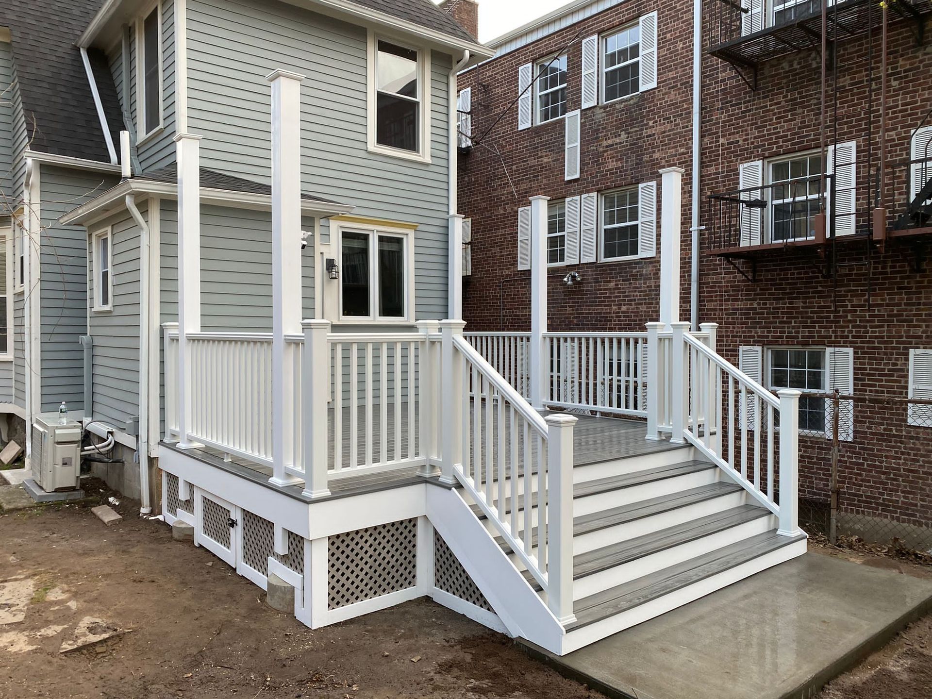 White deck with gray steps and railings, attached to a gray house, next to a red brick building.