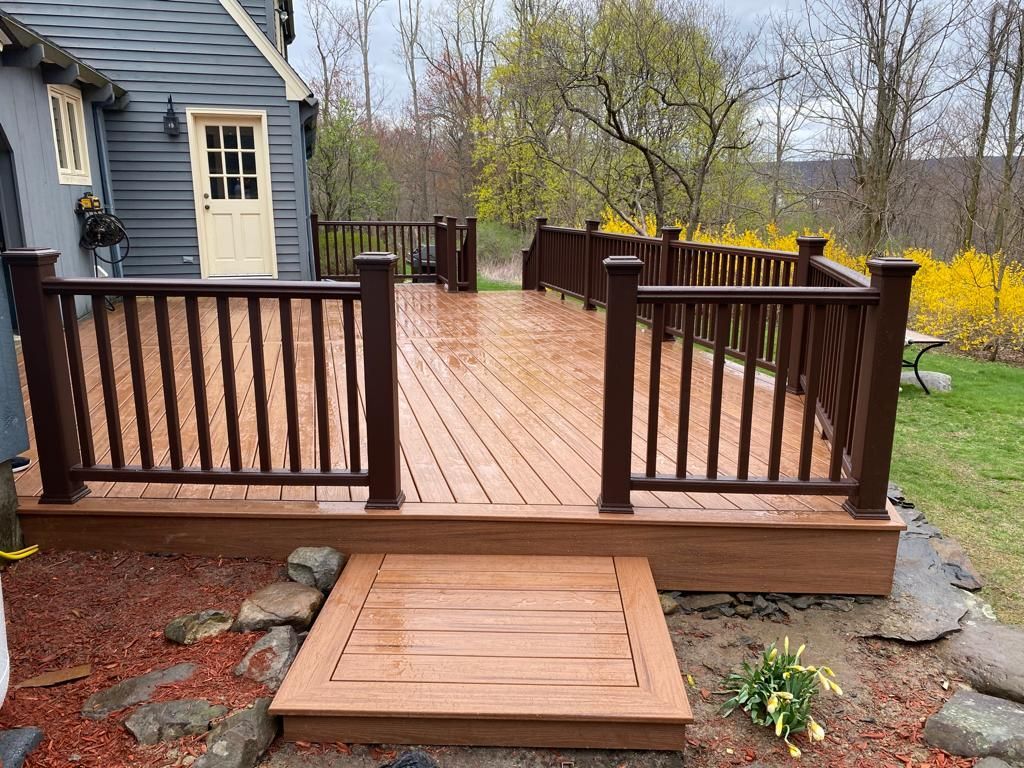 Brown wooden deck with railings, steps, and a door, attached to a gray house, set outside with plants and trees.