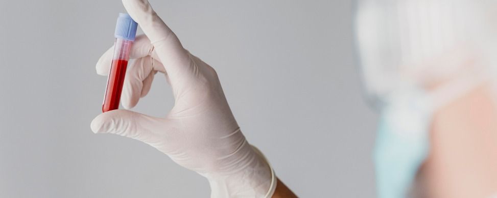 A gloved hand holds a blood sample test tube against a neutral background, with a blurred medical professional nearby.