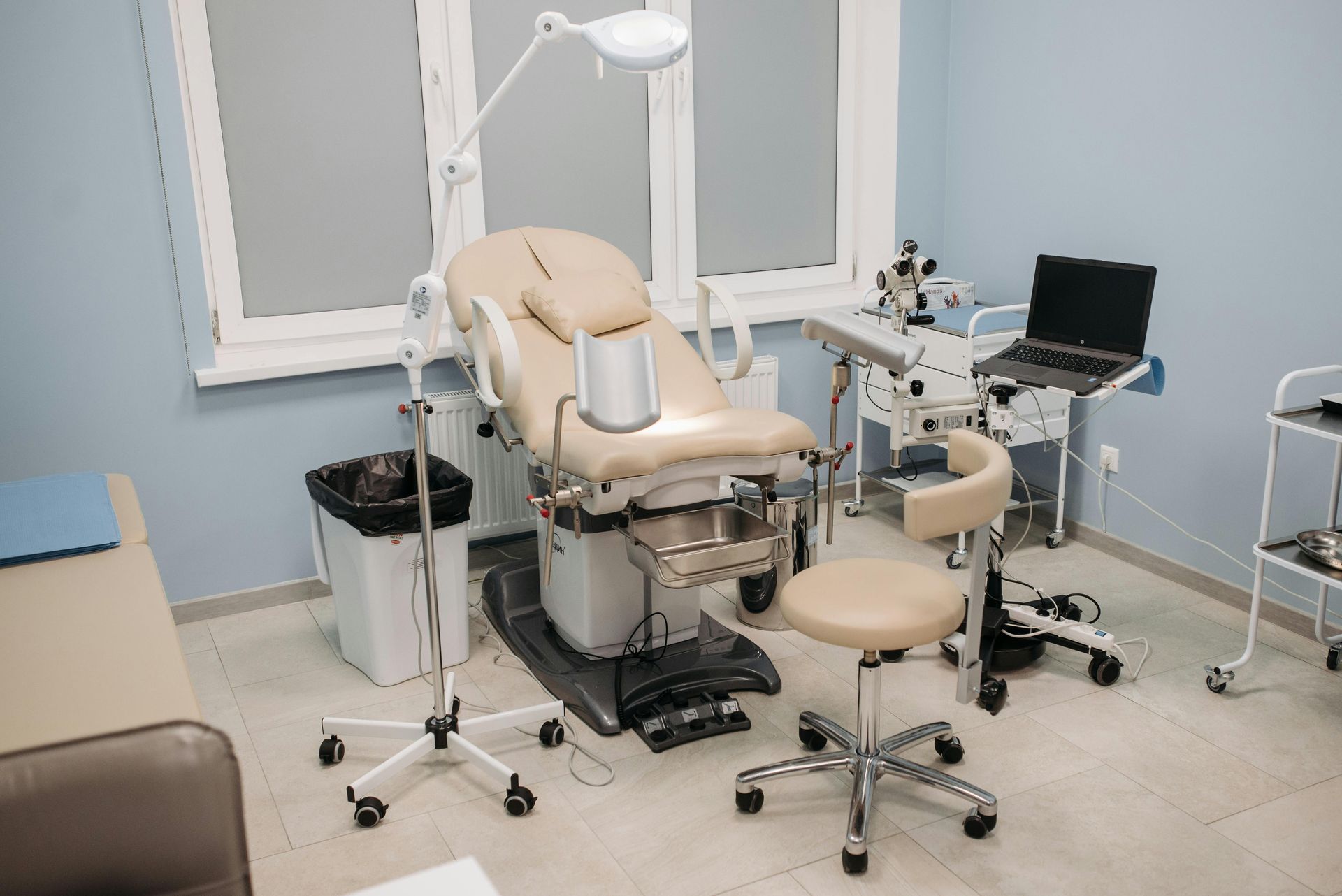 A medical examination room featuring a gynecological chair, a doctor's stool, diagnostic equipment, and a laptop.