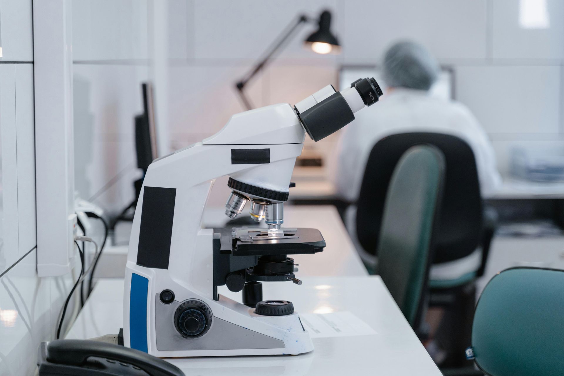 A white microscope sits on a laboratory table in the foreground, with a researcher working at a computer in the background.