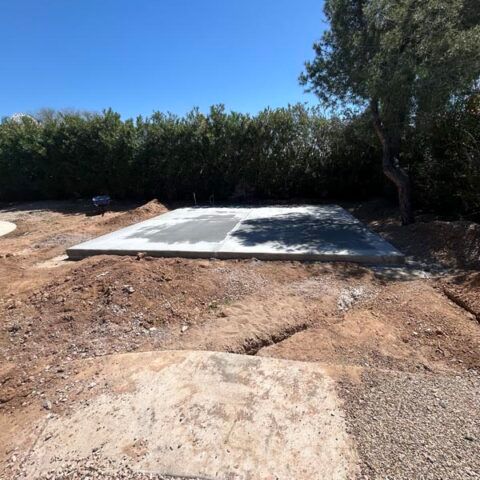 Concrete slab in a dirt clearing bordered by dense green bushes under a blue sky.