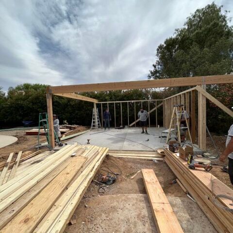 Partially framed house under construction with lumber stacks and workers on a concrete slab