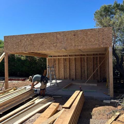 Wood-framed house under construction with lumber piles and two workers on a sunny day