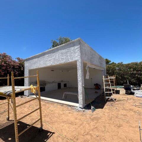 Small unfinished concrete building under construction on a sunny dirt lot with scaffolding nearby