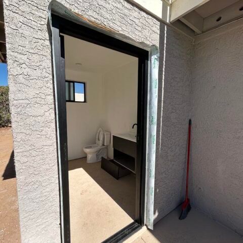 Open doorway to a small bathroom with toilet, sink, and dark vanity; broom leans outside on a patio.