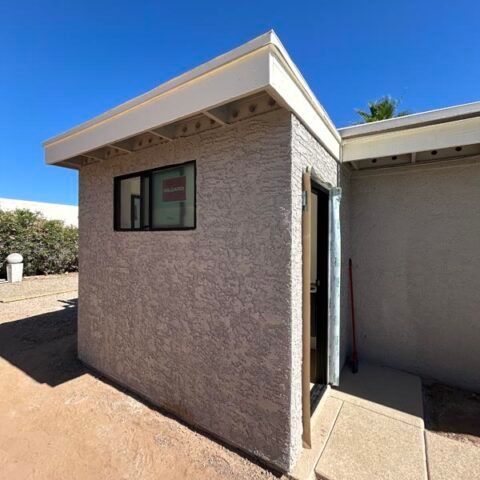 Small stucco building exterior with a window and open door in bright sunlight