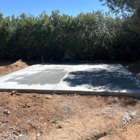Freshly poured concrete slab on bare ground beside dense trees under a clear sky