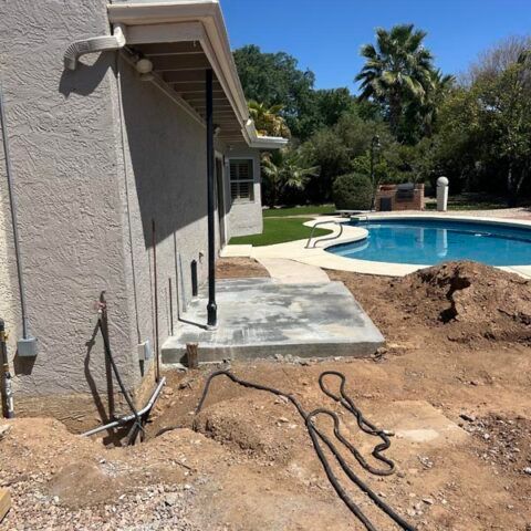 Backyard poolside patio with exposed dirt trench and utility cables beside a house