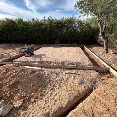A dirt foundation site with wooden form boards and a person working near the left side, trees in the background