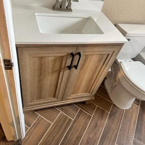 Bathroom vanity with white sink, wood cabinet doors, and herringbone tile floor beside a toilet