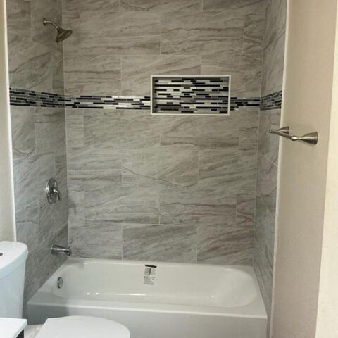 Small bathroom with gray tiled shower-tub combo and decorative mosaic accent strip.