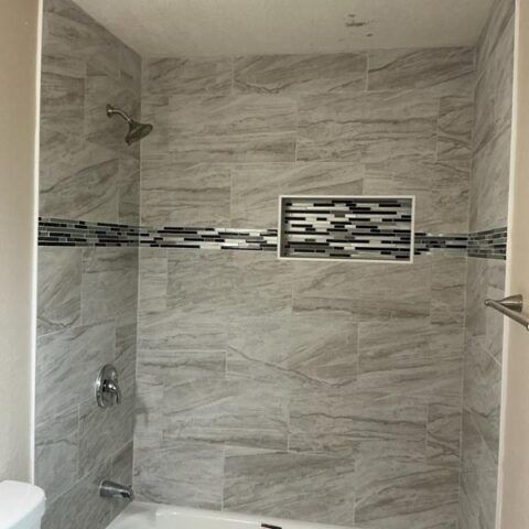 Modern tiled bathroom shower with gray stone-look walls and a decorative mosaic accent strip.