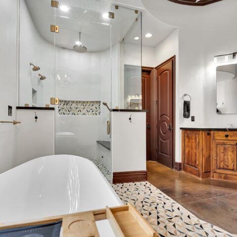 Bright bathroom with freestanding tub, glass shower, wooden vanity, and patterned tile floor