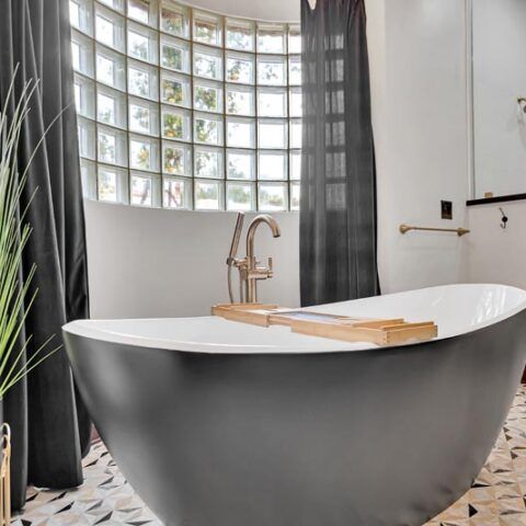 Modern freestanding bathtub in a bright bathroom with curved glass-block window and dark curtains