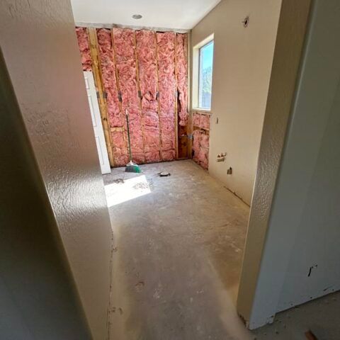 Empty unfinished room with pink insulation exposed on one wall and a small window at the back.