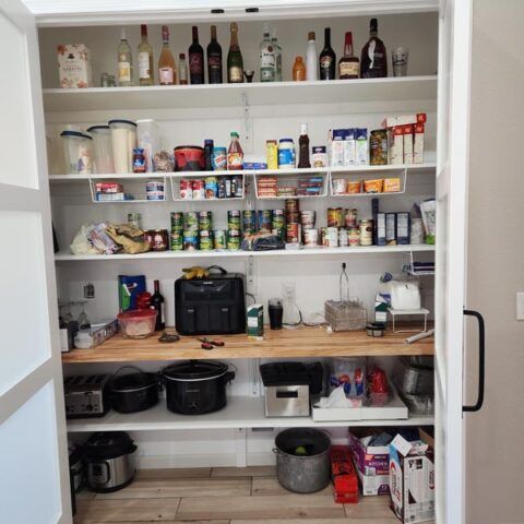 A pantry closet with shelves of food, bottles, appliances, and pots organized on a wooden counter.
