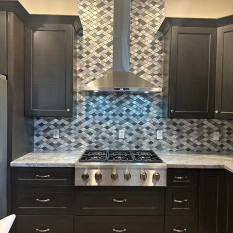 Dark modern kitchen with mosaic tile backsplash, stainless range hood, and gas stove under dark cabinets