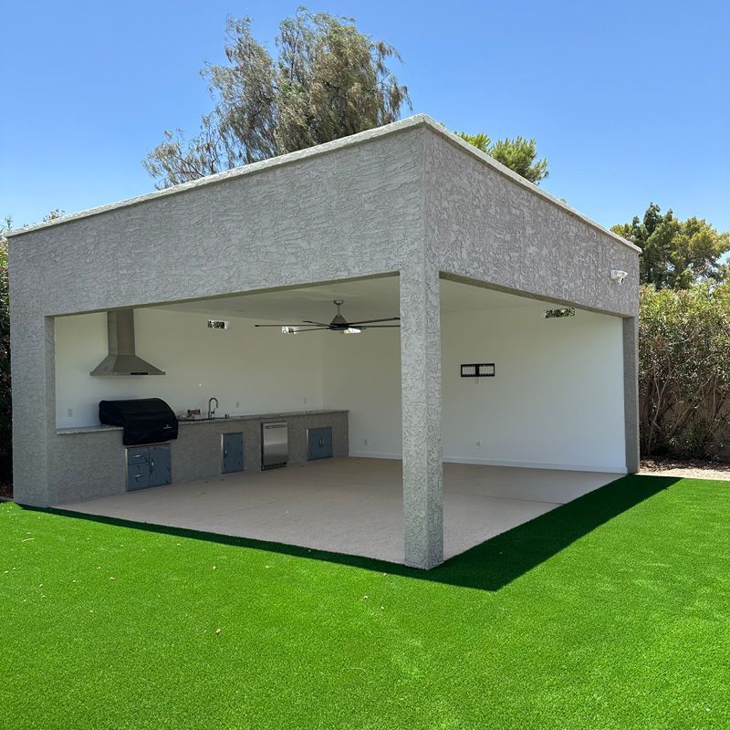 Covered outdoor patio with gray stucco roof, ceiling fan, grill, and bar counter over bright green turf