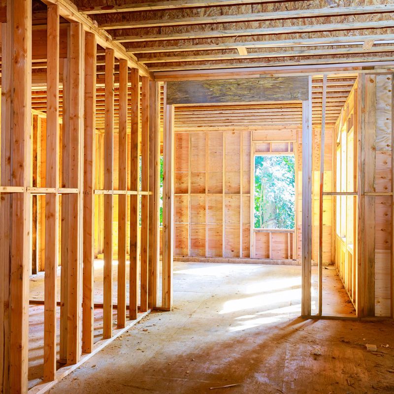 Unfinished wooden house interior with exposed framing, beams, and sunlight through open windows.
